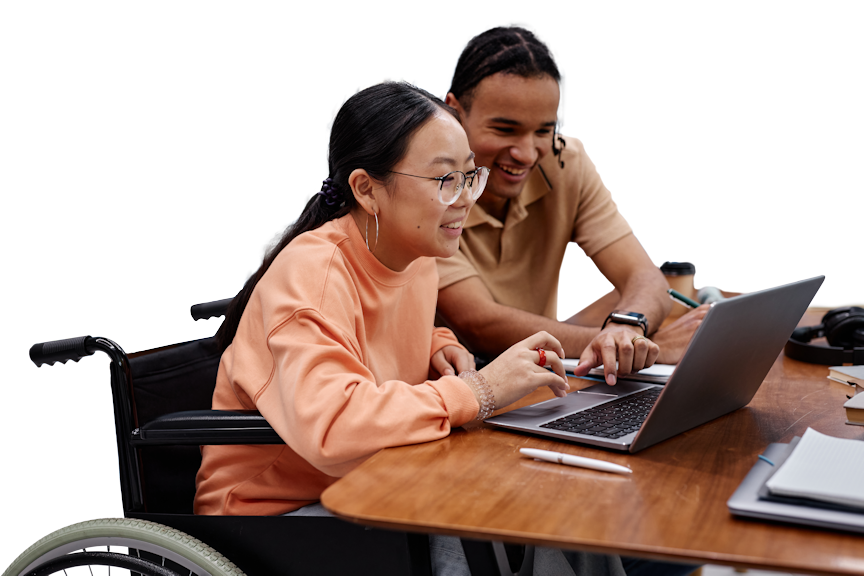 Zwei lächelnde Personen, die im Rollstuhl an einem Schreibtisch sitzend gemeinsam an einem Laptop arbeiten