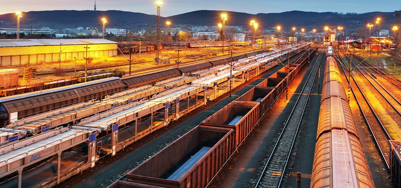 Güterzüge auf mehreren parallelen Bahngleisen eines Güterbahnhofs in der Dämmerung mit Beleuchtung vor einigen Hügeln in der Ferne.