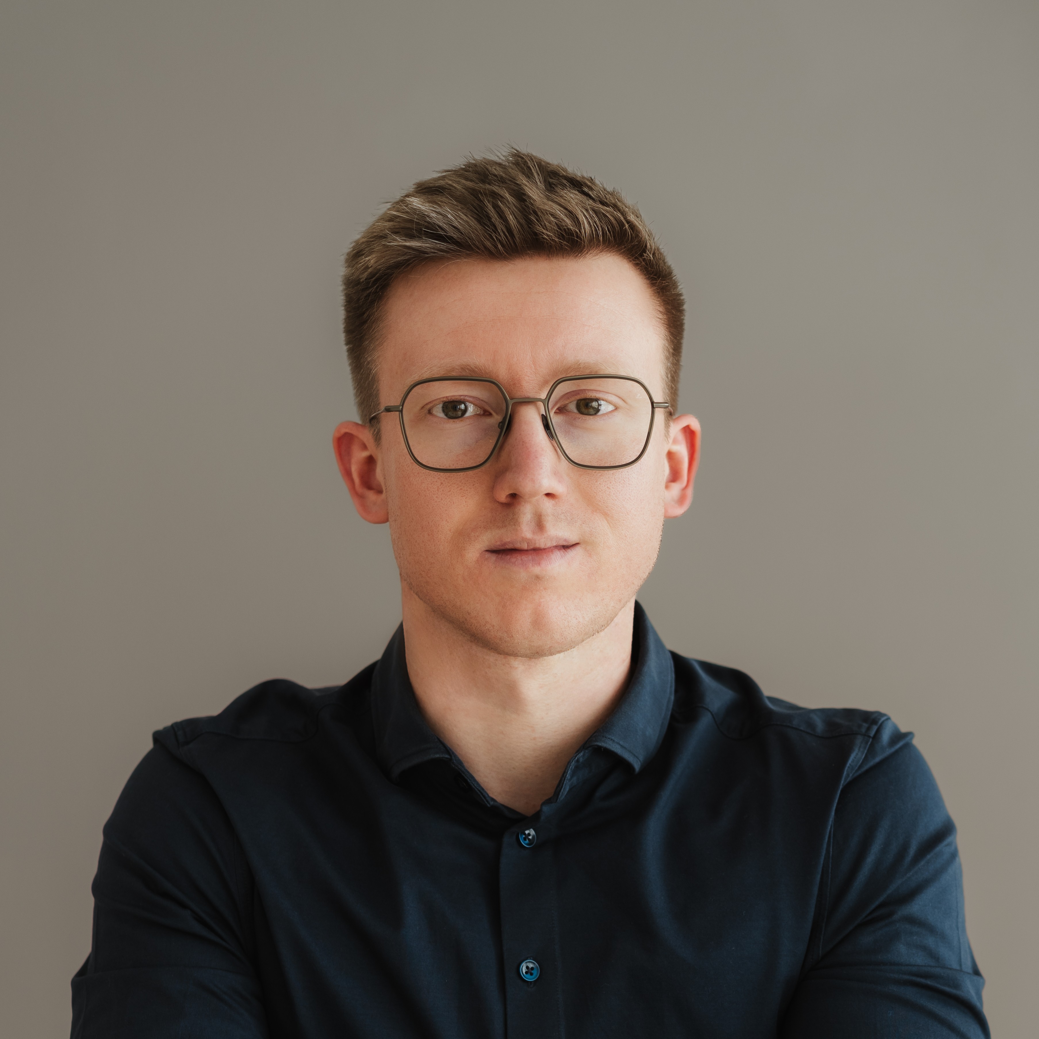 Porträtaufnahme einer Person mit runder Brille und dunkelblauem Hemd vor einem neutralen grauen Hintergrund. Die Person blickt direkt in die Kamera und ist mittig im Bild positioniert. Die Aufnahme ist gleichmäßig ausgeleuchtet und im Stil eines professionellen Business-Porträts gestaltet.