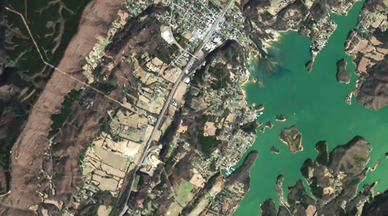 A satellite image of a coastal town where a river meets a body of water, surrounded by patches of green and cleared land