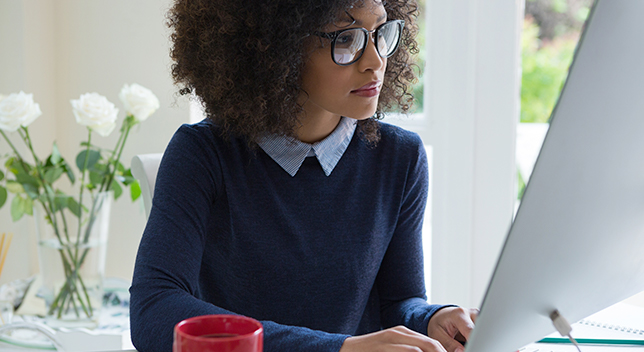 Junge Frau mit Brille an einem Schreibtisch, die auf einen großen silberfarbenen Computermonitor schaut